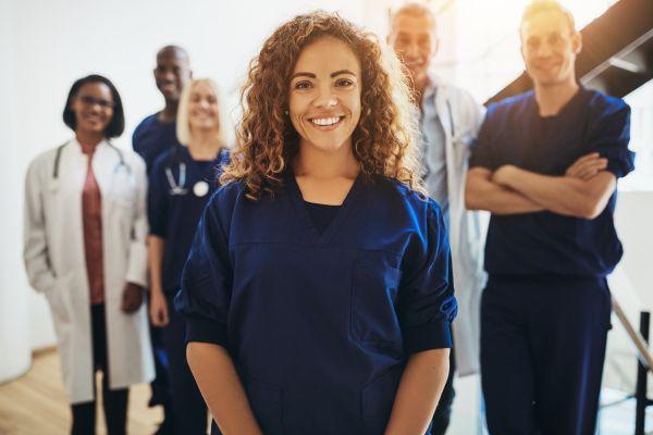 Happy woman in blue scrubs with a team of medical practitioners in the background.